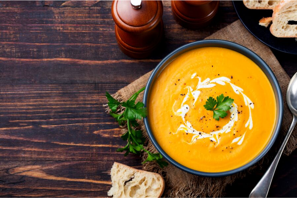 Sopa de calabaza servida en un tazón blanco, adornada con semillas, crema y trozos de chile. Al lado, calabazas decorativas y chiles.