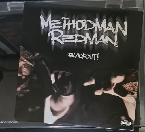 This is a photograph of a vinyl record album cover, resting inside a plastic sleeve. The cover is primarily black with two portraits of a person in a grayscale tone. The person is featured on both the left and right sides of the cover, looking towards the center. The person is wearing a bandanna and a tank top. The text on the cover reads "METHOD MAN" in large, white, stylized lettering at the top and “BLACKOUT!” below that in smaller lettering. A "Parental Advisory: Explicit Content" label is visible in the bottom right corner.