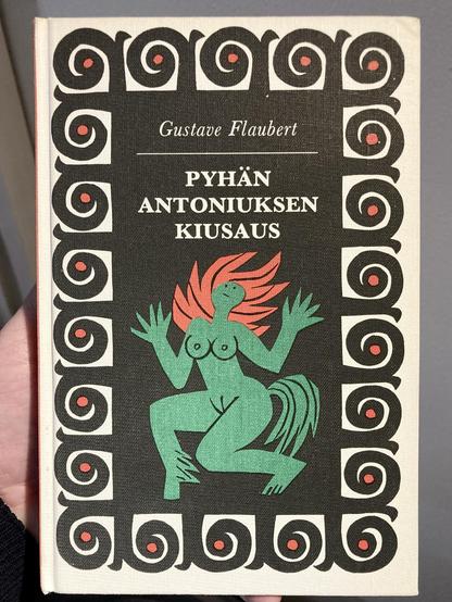 A book cover featuring the title "Pyhän Antoniuksen kiusaus" by Gustave Flaubert. The design includes a green figure with red hair and stylized features, surrounded by a decorative border of black spirals and red dots.