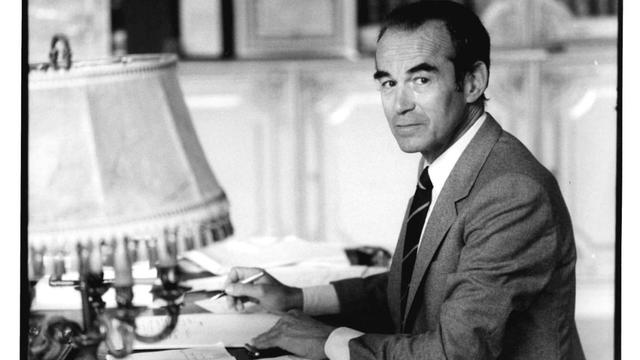 Photographie en noir et blanc de Robert Badinter, assis à son bureau du ministère de la Justice, tourné de 3/4. Archives nationales, cote 19950422/3