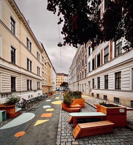 Foto durch die Gasse. Auf der Straße verteilt stehen unterschiedliche Straßenmöbel, die mit Bänken und Stühlen zum verweilen und klettern einladen und die mit integrierten Beeten die Straße begrünen.


Zusätzlich zur Möblierung lösen mit Farbe auf den Boden gehalte geometrische Formen in unterschiedlichen Farben den linearen Charakter der Straße auf und unterstützen somit den Platz ähnlichen Charakter