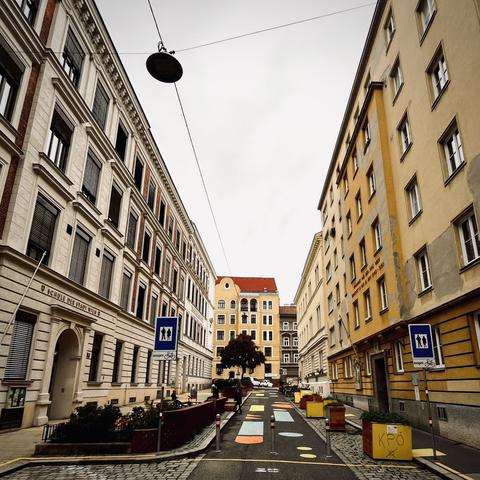 Die Straße von der anderen Seite. Im Bereich des Zugangs stehen Fußwegschilder mit einem Zusatzschild „ausgenommen Fahrräder“ - eine Reihe Poller unterstützt die Aussage der Beschilderung.