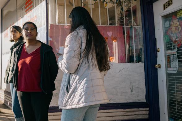 Pedestrians wait for the bus in a cluttered shop doorway.
