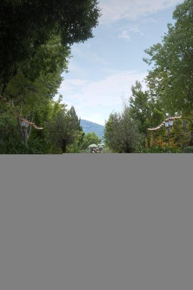 An artificial pond, probably a fountain, surrounded by trees and plants. Midway, 2 giants flower pots with small trees in them, at the far end, a statue and in the background, mountains, under a blue sky with some white clouds