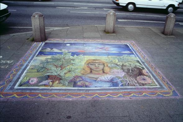 Foto im Querformat. Die Kamera blickt leicht nach unten auf den Gehsteig an einer Straßenkreuzung. Dort wurde ein prächtiges Bild mit Kreide gemalt, schätzungsweise 2 x 2 Meter oder größer. Es zeigt eine Person mit schulterlangen dichten gelbblonden Haaren zwischen grünen Pflanzen. Links und rechts neben der Person sind kleine dicke Bäume mit einem Gesicht auf dem Baumstamm. Im Hintergrund ist blaues Wasser, vielleicht ein See oder ein Fluß. Links und rechts hinter dem Wasser sind hellgrüne Berge. In der Mitte geht eine rote Sonne über dem Wasser auf oder unter.

Die Bürgersteig-Ecke wird durch Poller von der Fahrbahn abgegrenzt. Dort sind ansatzweise zwei Autos zu erkennen.