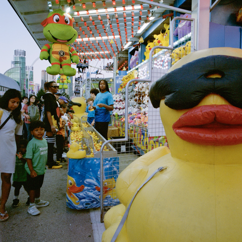 A giant inflatable yellow rubber duck wearing a black mask fills the left side of the frame a ninja turtles hang from the awning of a game involving floating rubber ducks.