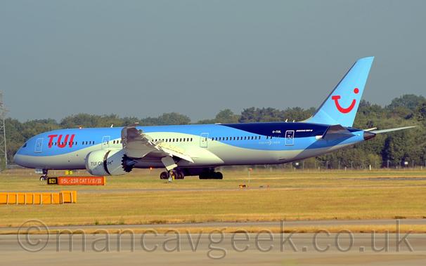 Side view of a twin engined jet airliner decelerating from right to left after landing.
The Plane is mostly light blue, with a creamy-white belly that swoops over the wings, and a dark blue section at the top of the rear fuselage.
There are large, red "TUI" titles on the upper forward fuselage, and the white registration "G-TUIL"on the upper rear fuselage, forard of the rear door, which is followed by white text "Boeing 787-9".
There are cream-coloured "DreamLiner" titles on the lower rear fuselage.
The tail is a pale blue, with a red logo that is somewhere between the lowercase letters "t", "u", and "I", and a red winkey face.
The engine pods are a creamy colour, with the black web address "tui.com" on the side.
The front and rear halves of the engine pods are split, allowing the thrust reversers to work.
Green grass fills the foreground, with trees lining the frame in the background, under flat grey sky.