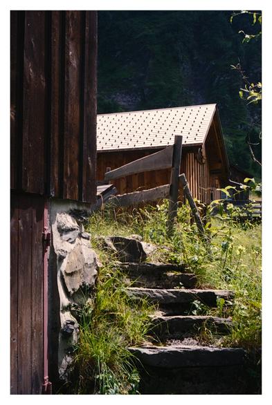 Color photograph of a rustic stone staircase leading through overgrown grass to a wooden fence and a traditional wooden hut with a tiled roof, nestled in the green, mountainous landscape of the Tschinglenalp.