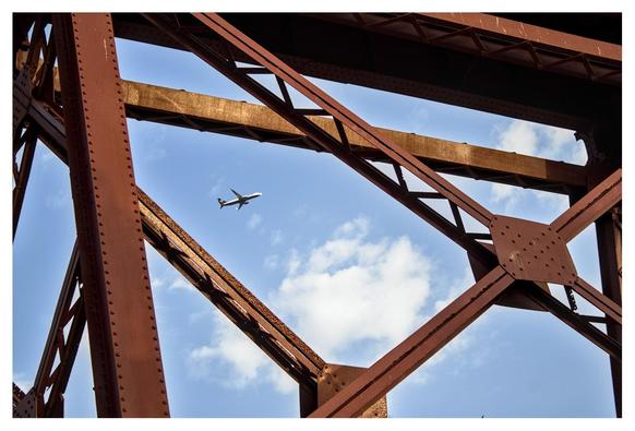 The image shows the iron supports of a red bridge (the so-called 'Cable Inglés' in Almería, a city in southern Spain), and through them, a plane can be seen flying across the sky among the clouds.
---
La imagen muestra los soportes de hierro de un puente de color rojo (el llamado ‘Cable Inglés’ de Almería) y, a través de ellos, se ve un avión surcando el cielo entre las nubes.