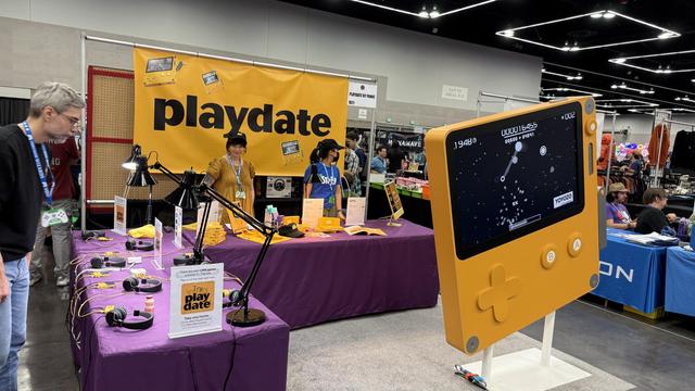 The Playdate booth from last year's Portland Retro Gaming Expo. A large yellow Playdate in front of two purple tables with merch and Playdate consoles!