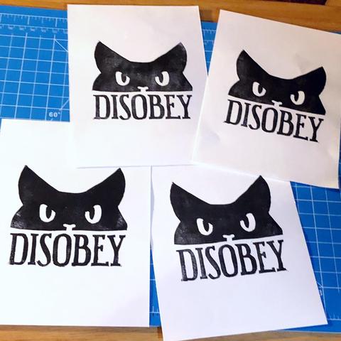 four block prints on white paper laid out messily on a blue cutting mat. the prints are of the top half of a black cat's head looking very grumpy and the word "DISOBEY" is block caps underneath.