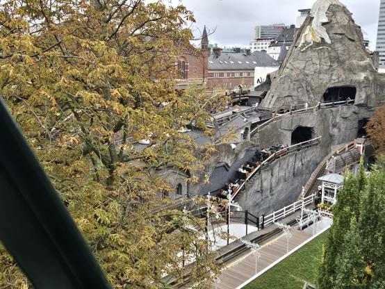 View from above of Tivoli's Rutschebanen. An imposing fake mountain peeks out from behind fall foliage, with wooden coaster tracks running along dipping ridges in the mountain and into and out of dark holes. A coaster train is ascending one of the dips, with passengers and a brake operator behind the lead car.