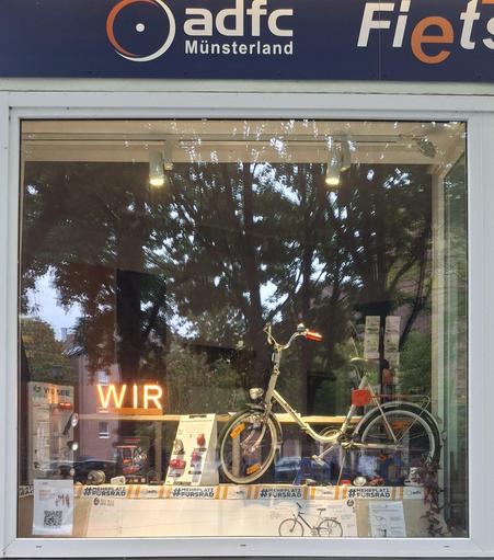 Schaufenster der ADFC-Münsterland Geschäftsstelle "Fietserei". Im Schaufenster steht rechts ein Klapprad der 70er Jahre ausgestattet mit der damaligen Fahrradelektrik und Reflektoren sowie einer Abstandskelle. 
In der Mitte steht ein Fahrradlicht-Display der 90er Jahre.
Links steht ein Fahrradlicht-Display der 2010er Jahre.
Oberhalb der Displays leichtet ein orangenesc"WIR".
Über dem Schaufenster steht auf blauem Grund in weißer Schrift "ADFC-Münsterland " mit dem Reifenlogo des ADFCs.
Bäume spiegeln sich in der Fensterscheibe.