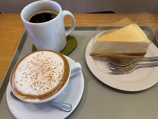 A grey tray with a cup of coffee, a cappuccino, and a slice of cheesecake.