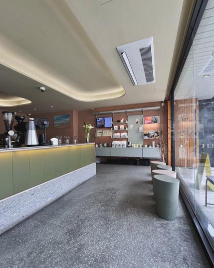 A café interior with a coffee counter, display shelves, and seating area. The space features a terrazzo floor, olive-green accents, and a large window.
