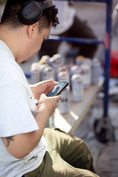 A spraycan artist sitting on a scaffolding, looking at his phone. In the blurry background, we can see an army of spray cans.