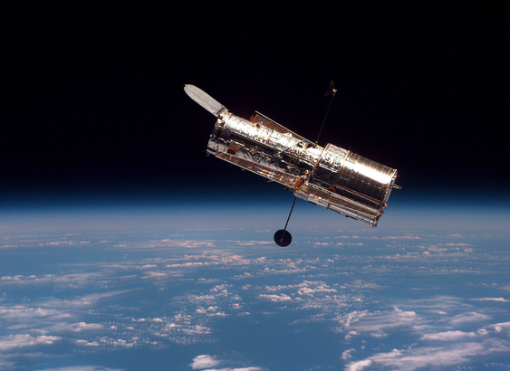 The Hubble Space Telescope orbits above Earth,. Below, the curve of Earth is visible with a blue atmosphere fading into darkness and white clouds scattered across the surface.