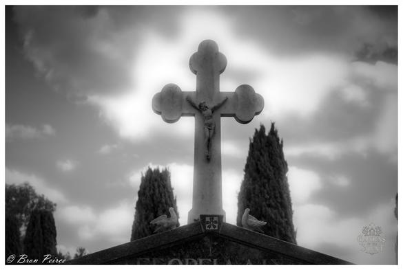 A dramatic, black and white, low angle photograph of a crucifix atop a headstone, signed by Bron Peirce.

The marble cross, featuring the figure of Christ (corpus), stands silhouetted against a bright, cloudy sky, with light breaking through the clouds behind it.

Below the cross, two small dove sculptures rest on the tombstone's top edge. Two tall, dark cypress trees flank the cross, drawing the eye upward. The image has a solemn and ethereal quality.