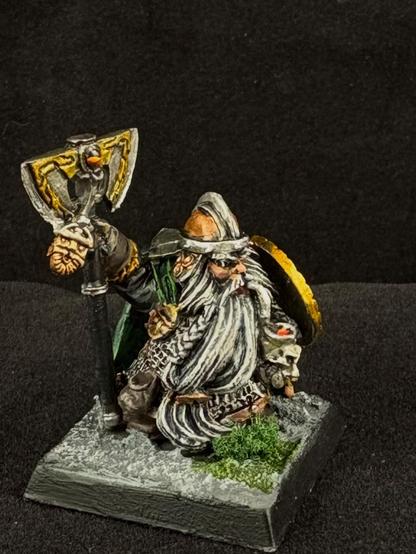 A Warhammer dwarf miniature with a long grey beard leans against a large two-handed axe while smoking a pipe made from a goblin skull.
