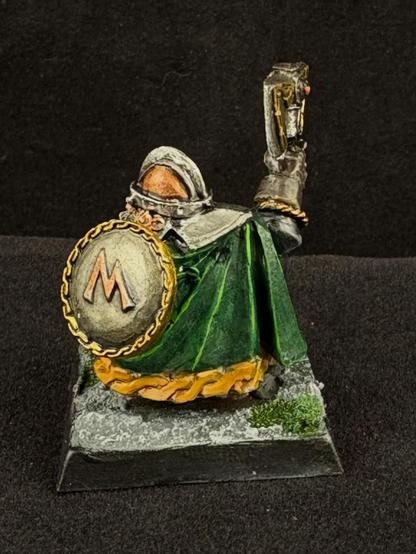 The back of the dwarf miniature. He wears a copper helmet and a long green cloak trimmed in yellow and carries an iron shield decorated in gold.