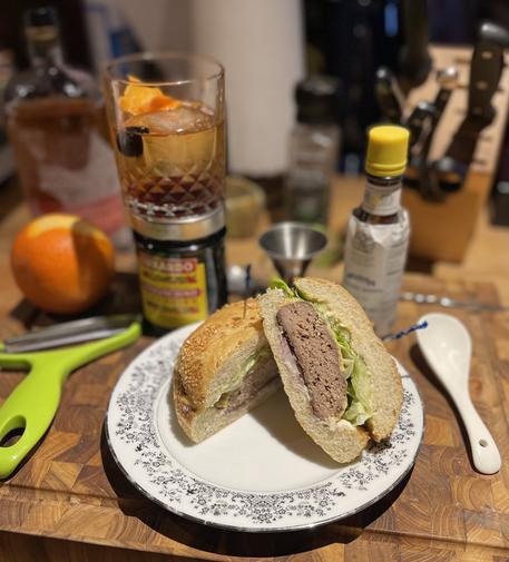 a cheeseburger cut in half and arranged on a plate. there’s ingredients and a mixed old fashioned behind it.