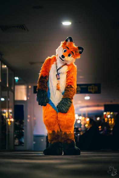 An orange fox fursuiter (ToasterTheFox) standing in the middle of a hotel lobby with his head tilted to the side. The photo is taken from below.