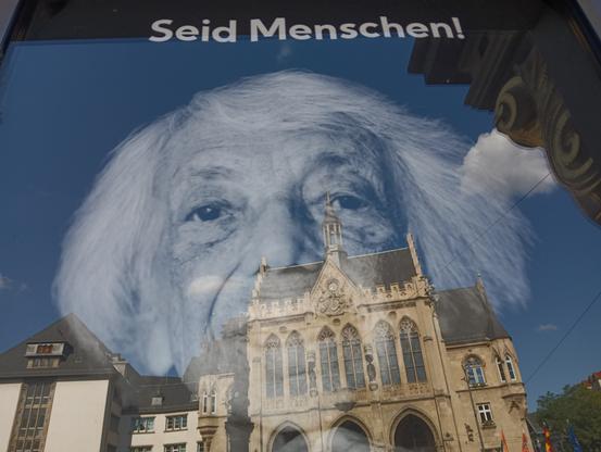 Foto eines Plakats das hinter einer Glasscheibe mit Spiegelung hängt. 

Das Plakat zeigt den weißhaarigen Kopf der Margot Friedländer, darüber steht: SEID MENSCHEN! 
In der Scheibe spiegelt sich das neugotische Erfurter Rathaus unter blauem Himmel. 
Übrigens: Gesehen neben dem Eingang der Kunsthalle Erfurt (September 2025).