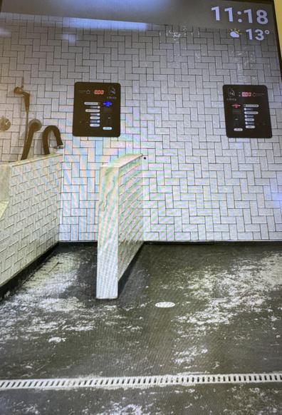 The image shows a bathroom or shower area with white tiled walls, a floor drain, and a foot-operated faucet. Wall-mounted digital panels display water settings. The environment appears clean but somewhat worn.

Das Bild zeigt einen Badezimmer- oder Duschbereich mit weißen Fliesenwänden, einem Bodenablauf und einem Fußwasserhahn. An der Wand montierte digitale Paneele zeigen Wassereinstellungen an. Die Umgebung wirkt sauber, aber etwas abgenutzt.