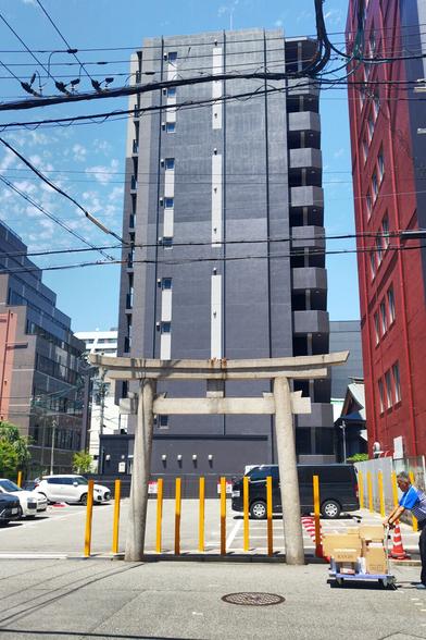 It's a stone gate that stands isolated at the side of a parking lot. There are medium rise buildings in the background and a delivery man is pushing into the shot with a hand cart from the right. Only very thin people will be able to walk through this gate because they have erected quite closely to each other tall yellow poles right behind it. This gate makes limited sense in this location. There is a shrine nearby and I'm guessing this used to be on the path to the shrine before this became prized real estate.