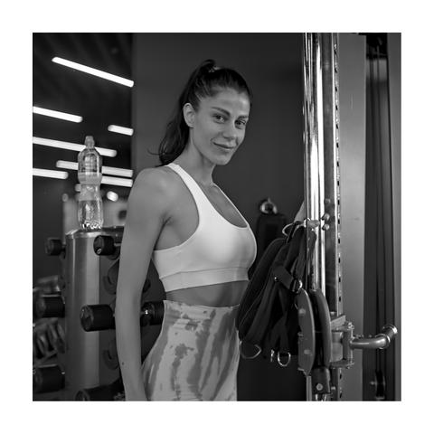 A square, black and white portrait of a woman standing next to a chrome weight rack in a gym. She is looking at the camera with a gentle smile, wearing a white sports bra and tie-dye leggings. A water bottle rests on the equipment to her left, and a dark bag hangs from a vertical bar on the right.