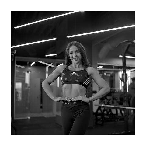 A square, black and white portrait taken in a gym. A fit woman with long hair stands smiling directly at the camera, posing with her hands on her hips. She is wearing a patterned Adidas sports bra and dark leggings. The background is dark with bright, linear overhead lights.