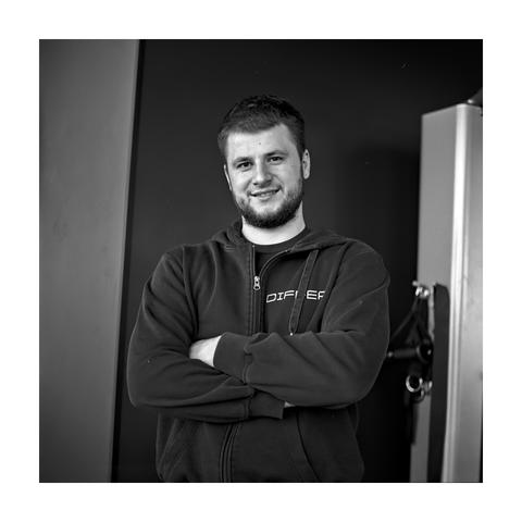 A square, black and white portrait of a man with a beard and short hair, standing in a gym with his arms crossed. He is smiling slightly while wearing a dark, zippered hoodie. The background is simple and dark, with a piece of light-colored exercise equipment visible on the right