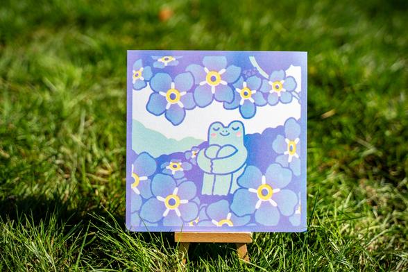 Photograph of a square risograph print of a frog chilling in a field of blue flowers. The frog is sitting down with his arms holding his legs, he looks very calm with his eyes closed and a smile on his face. The print is rested on a small standee that is standing in a field of grass.