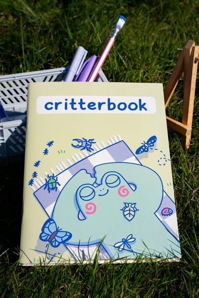 Photograph of a stickerbook with the title "critterbook" showing a frog laying down with his arms behind his head on a blanket surrounded by bugs and butterflies. The main colour of the book is a yellowish green. The book is rested on a blue crate with pens in it standing in a field of grass.