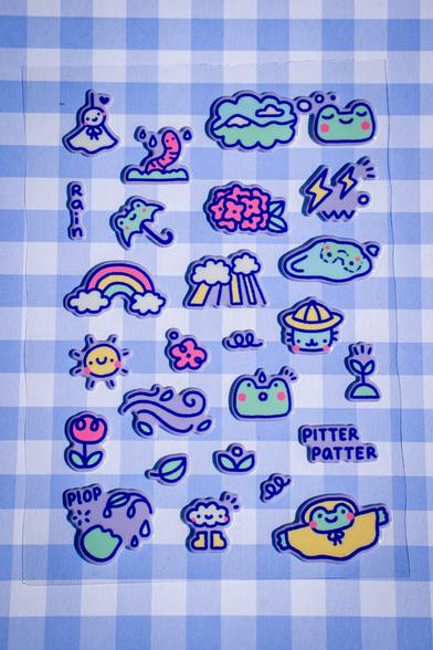 Photograph of a stickersheet. The stickers are puffy so a bit 3D and show a variety of small rain themed icons: A good weather ghost, a worm, a frog dreaming of the mountains, the word "rain", an umbrella shaped like a frog, some red hydrangea, some lightning, a rainbow ending in two clouds, sun coming through the clouds, a puddle showing the reflection of a frog, a smiling sun, a red flower, a blue cat in a yellow rain hat, another flower, the wind (just swirly lines), a frog looking surprised at a rain drop, a sprout, a leaf, the words "pitter patter", a frog hand reaching towards dew on a leaf with the word "plop", a cloud with rain boots smiling and a frog in a raincoat. The background ist a purple-white gingham.