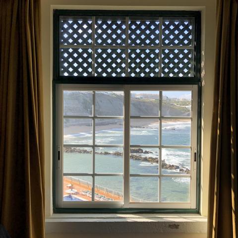 Through a paned window, a sunlit coastline beneath a steep cliff face can be seen. In the view’s foreground, a large natural rock pool seems to invite swimmers, and a row of deck chairs on a hotel deck can just be made out in the bottom corner. Inside, heavy curtains have been drawn to the side, and a lattice over the top third of the window helps reduce the glare from outside.