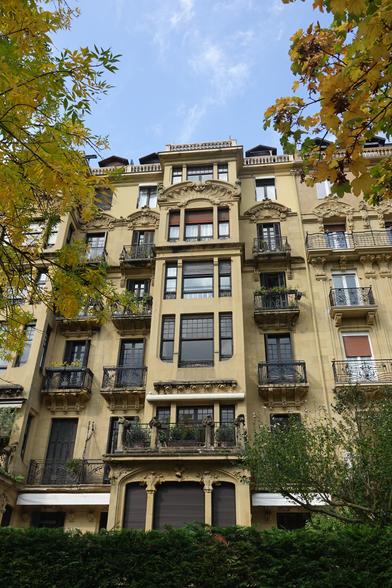 La façade d'un immeuble de style Art Nouveau.De grandes baies vitrées, de formes diverses ( rectangulaires, aux coins arrondis), surplombées par des chapiteaux ouvragés. La terrasse sur le toit est délimitée par un garde corps en pierre.