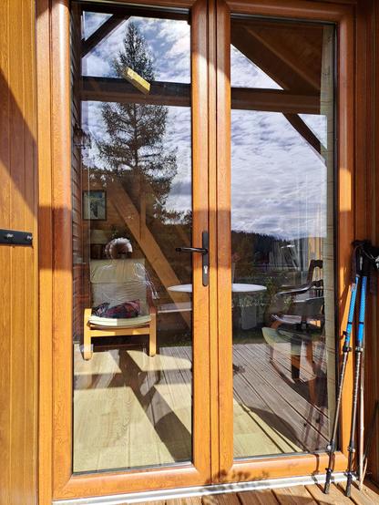 Une porte-fenêtre en bois, vue de l'extérieur. Le paysage se reflète dedans. Ciel un peu chargé, arbre, table blanche, fauteuil de jardin vert. On voit une personne assise dont on doute qu'elle soit dehors ou dedans, sur un fauteuil en tissu clair.