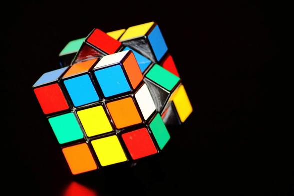 Le fameux casse-tête des années 80 : Le Rubik's Cube.
Sur fond noir, il est repose sur un coin. Pas une seule face est de la même couleur. Les faces visibles présentent des carrés de couleurs vives — rouge, bleu, vert, jaune, orange et blanc — formant un contraste saisissant avec le fond noir.
La lumière éclaire le cube en diagonale, mettant en relief ses arêtes brillantes et ses surfaces légèrement réfléchissantes.
L’ensemble évoque à la fois le jeu, la logique et la complexité d’une énigme à résoudre. Un jeu de mémoire ?