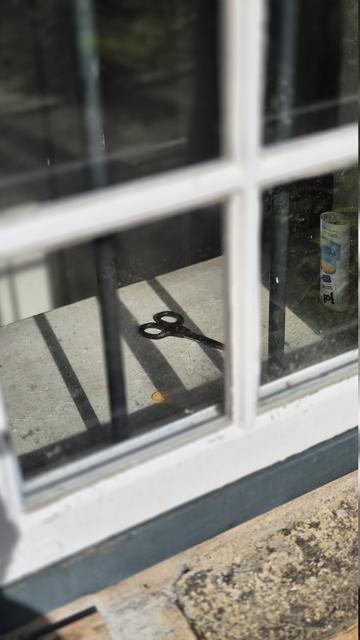 Nahaufnahme eines Teils eines weißen Sprossenfensters. Die Aufnahme erfolgte im Außenbereich. Der Blick geht dabei von oben links nach unten rechts durch die Fensterscheiben in das Innere eines Raumes. Zu sehen ist ein Fensterbrett, auf dem eine Schere liegt. Rechts davon steht eine Dose. Unmittelbar hinter der Fensterscheibe scheinen zwei senkrecht verlaufende, runde Metallstäbe mit Abstand zueinander zu verlaufen.