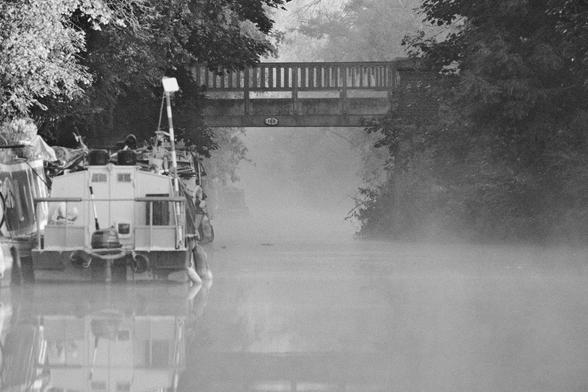 Very foggy view of a canal going under a low bridge with a boar on the left.