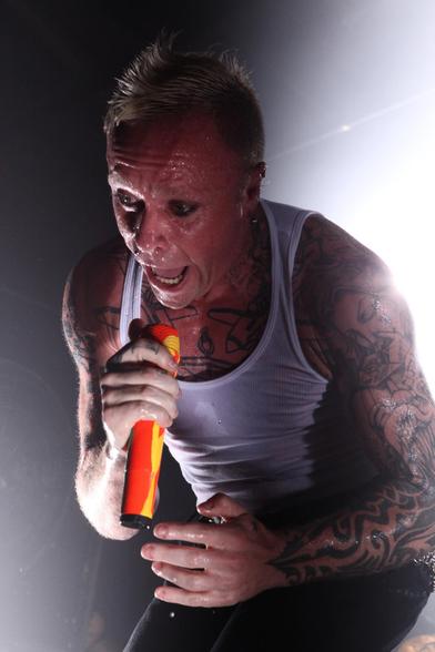 Keith Flint - a man with a short blonde mohawk hairstyle wearing a white vest (UK style) top sings into a microphone.