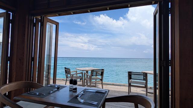 A wide angle view through folding wooden doors that make the floor to ceiling window looking over a turquoise blue sea with soft white clouds in the blue sky. Inside the window is a table set for two and outside there are tables with chairs for dinner on the wooden deck because it's too hot to eat in the midday sun.
