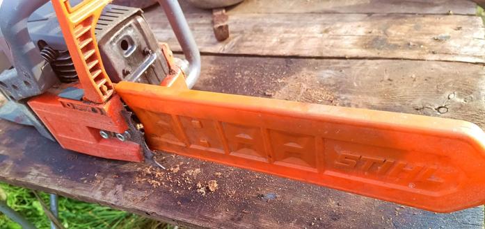 A chainsaw with dark grey and orange body. The bar is covered by a mismatching orange Stihl plastic protective cover. The sun shines through the plastic, revealing the bar inside being somewhat shorter.