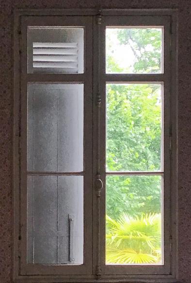Une fenêtre fermée. À gauche un volet est fermé. À droite feuillage vert clair des arbres dans le soleil