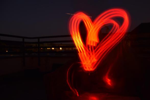 Das Foto wurde draußen im Dunkeln aufgenommen, es handelt sich um eine Lichtfotografie. Im Hintergrund sind sehr schlecht Lichter, eine Häusersilhouette und ein Balkongeländer zu erkennen. Das Bild wird bestimmt durch ein großes, etwas wackeliges und verwischtes Herz in Rottönen, dass in die Dunkelheit "gemalt" wurde.