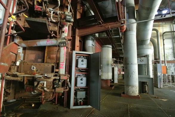 Aufnahme eines Inneren einer Fabrik. Rostige Leitungen, grosse Röhren und ein grosser Schaltschrank machen dies deutlich.