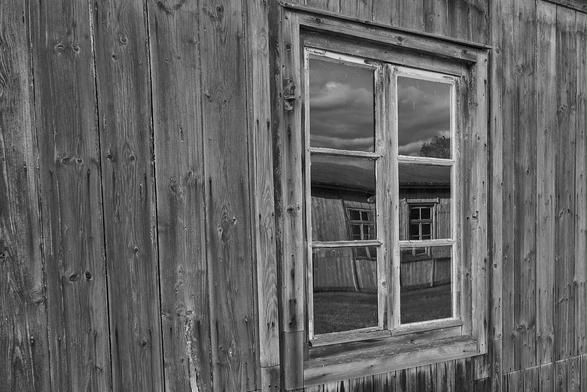 Ein Fenster einer Holzbaracke. Das Fensterglas ist sehr alt und damit noch von Hand gemacht. Somit ist die Oberfläche leicht wellig. Im Fenster spiegelt sich eine Nachbarbaracke und die Spiegelung ist - wg. der unebenen Glasoberfläche - leicht verzerrt.