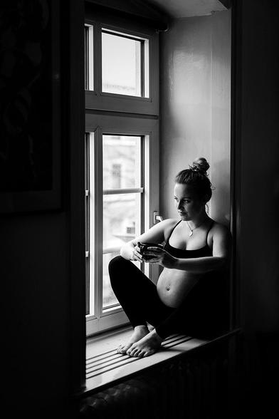 Eine schwangere Frau sitzt barfuß auf der Fensterbank in einem Fensterrahmen. Sie trägt eine schwarze Sport-BH und schwarze Leggings. Ihre Beine sind angewinkelt, die Füße ruhen auf der Fensterbank. Sie hält eine Tasse mit beiden Händen vor ihrem Bauch. Ihr Haar ist zu einem Dutt zusammengebunden. Das Bild ist schwarz-weiß und zeigt eine ruhige, nachdenkliche Atmosphäre.