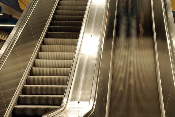 Zwei nebeneinander liegende Rolltreppen. Die eine steht still, die andere bewegt sich. Durch die Langzeitbelichtung ist die Bewegung verschwommen und man sieht schemenhaft eine Person.